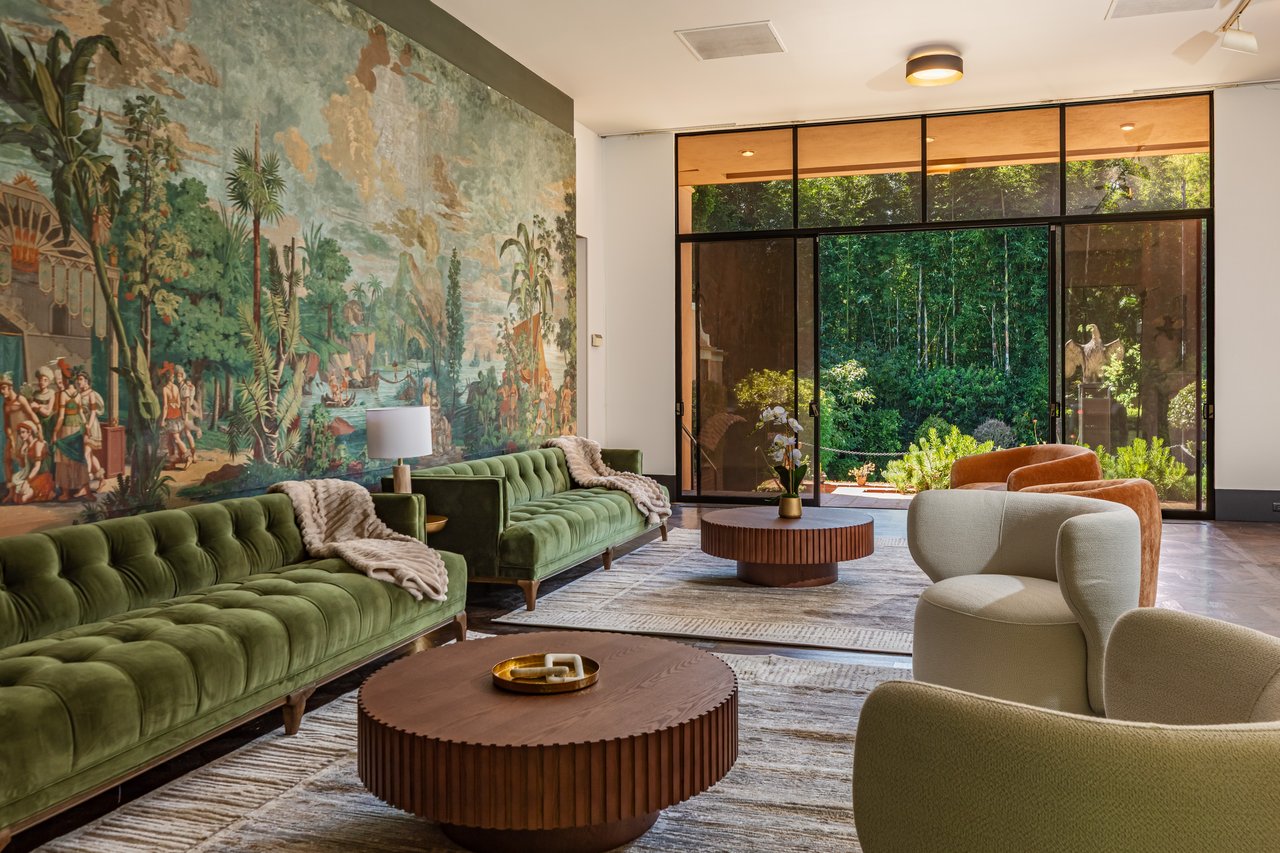 Living room with green velvet sofas, a large wall mural, and glass doors overlooking a lush garden.