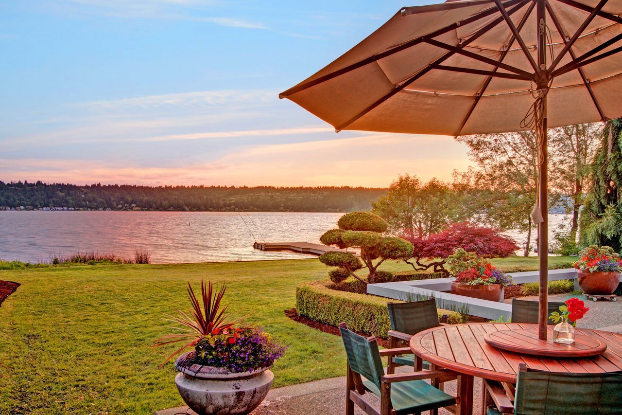 A patio with a table, chairs, and umbrella overlooking a lake at sunset