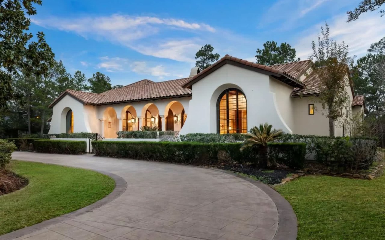  A luxurious Mediterranean-style house with a light-colored stucco exterior and terracotta tile roof is set amidst a lush green landscape.