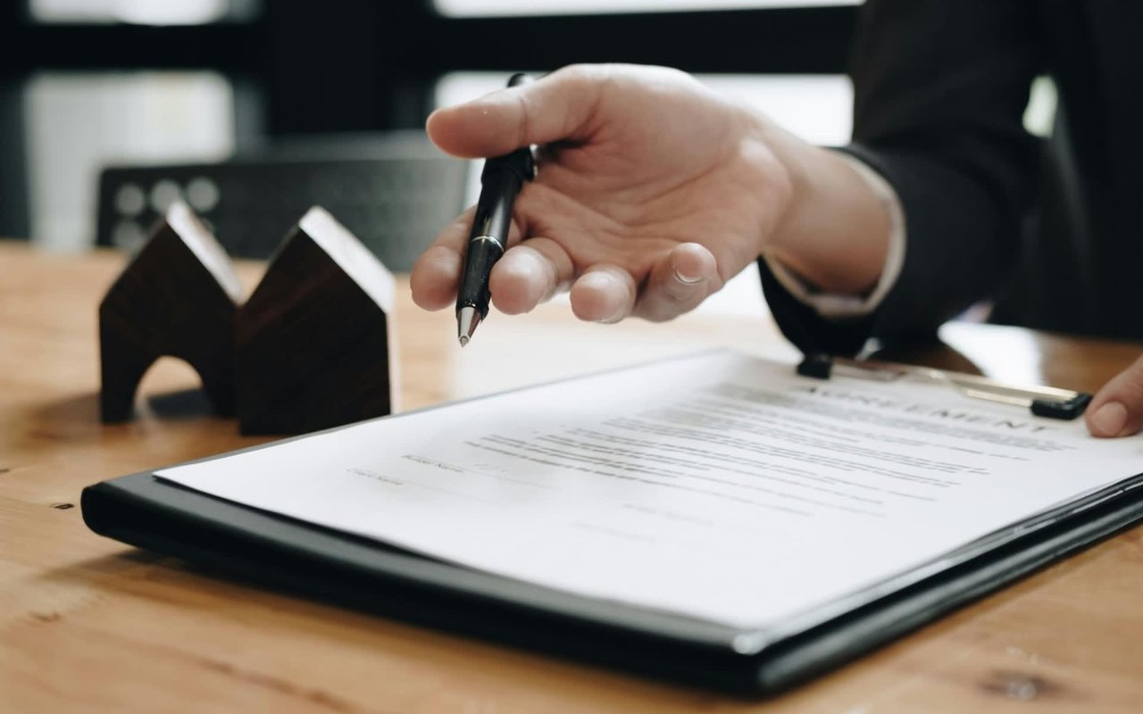 A hand gestures towards a clipboard with a legal document and a house model.