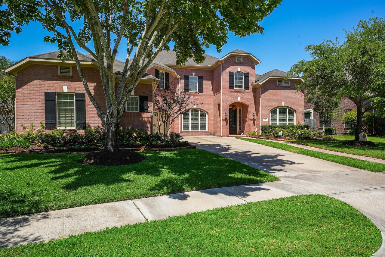 A  two-story Colonial-style red brick house with a lush front lawn, white trim, black shutters, and a driveway.