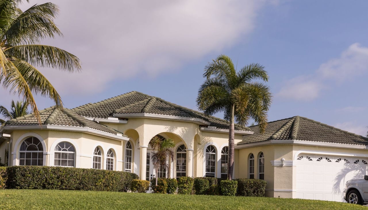 A grand, cream-colored house with a dark green tile roof stands on a well-manicured green lawn under a partly cloudy sky. 
