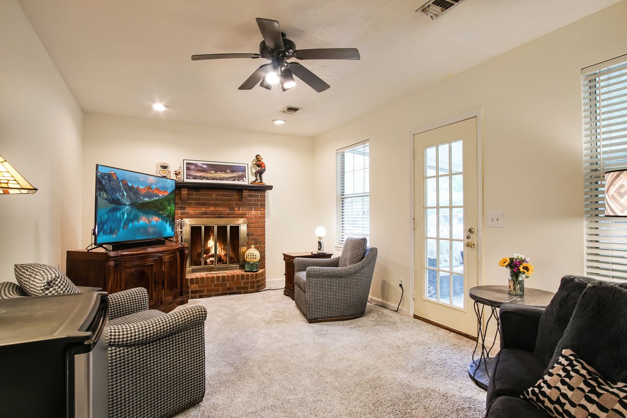 A cozy living room with comfortable seating, including chairs and a sofa. The room features a fireplace, a ceiling fan, and large windows allowing natural light to enter.