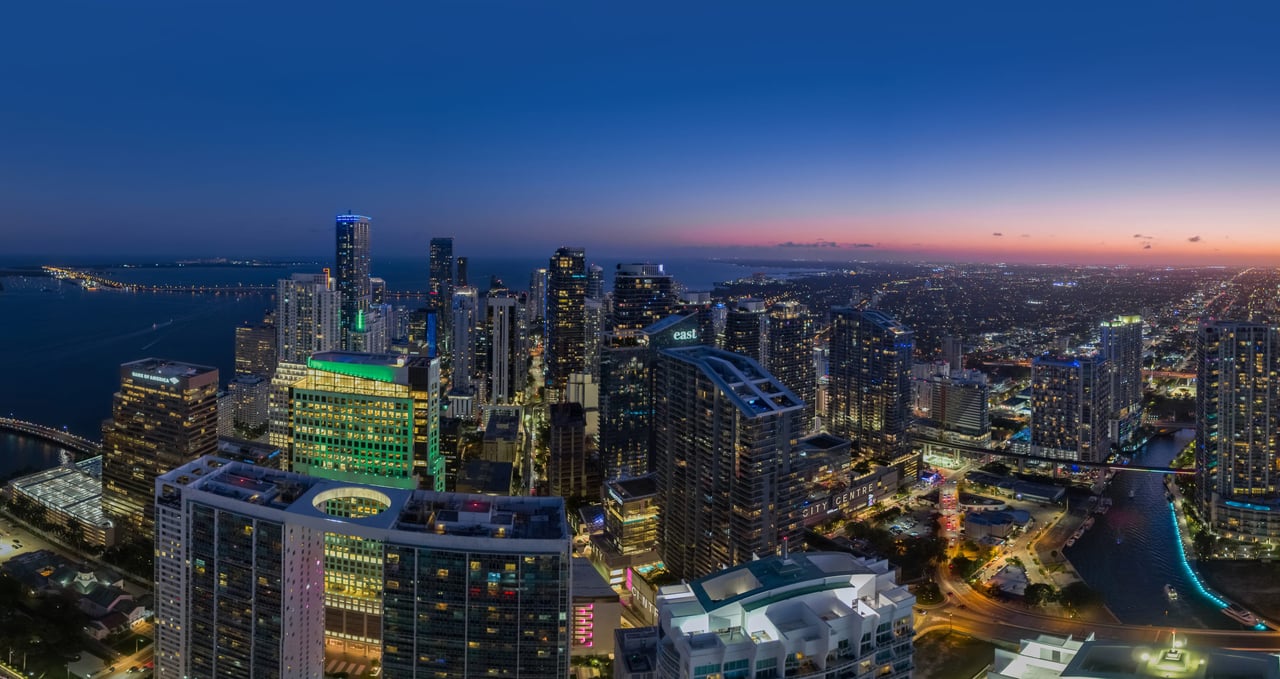Downtown skyline at dusk, with illuminated towers, a river, and city lights beneath a blue and pink sky.