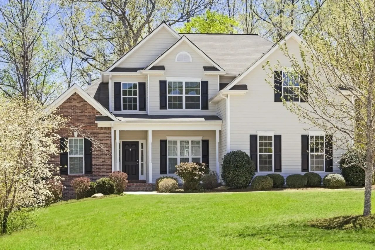  two-story white house with black shutters and a brick facade, surrounded by green trees and a manicured lawn.