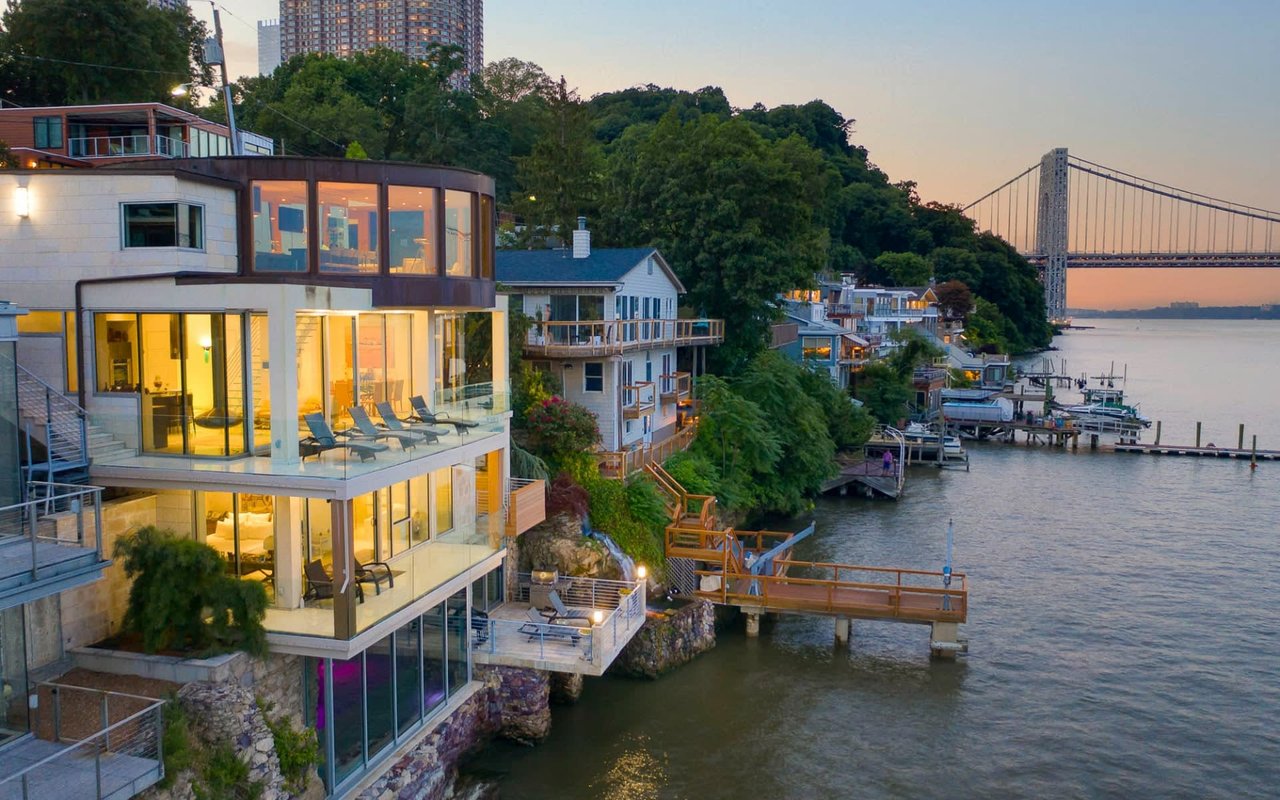 A row of diverse houses by a calm river with balconies, a rustic dock, and surrounded by lush trees.