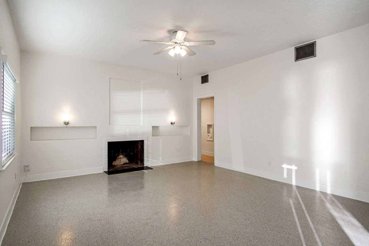 A living room featuring a fireplace, ceiling fan, and large windows. The room is spacious and well-lit with tiled flooring.