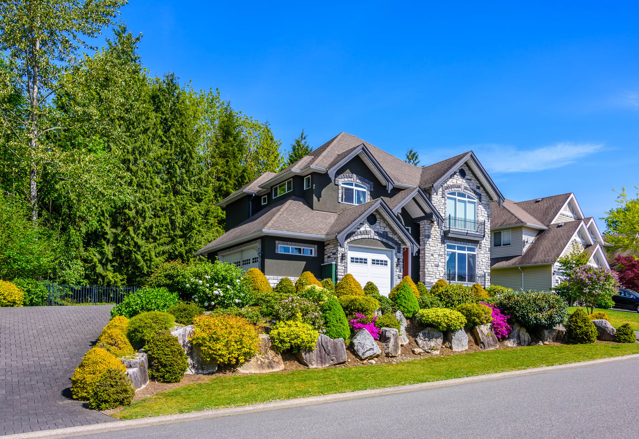 A large house with a lot of flowers in front of it. The house has a brown roof and white siding.