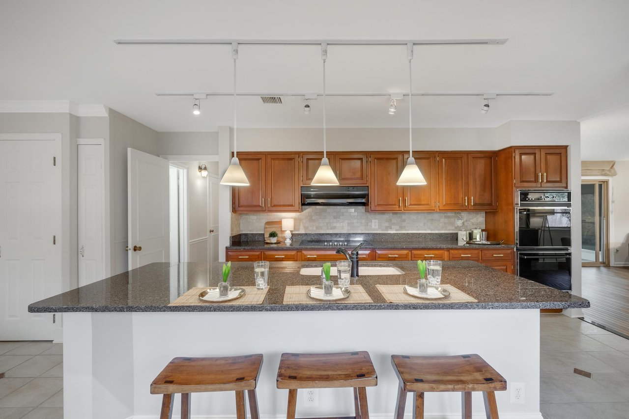 Kitchen with warm wood cabinets, double ovens, and a large granite island with three stools.