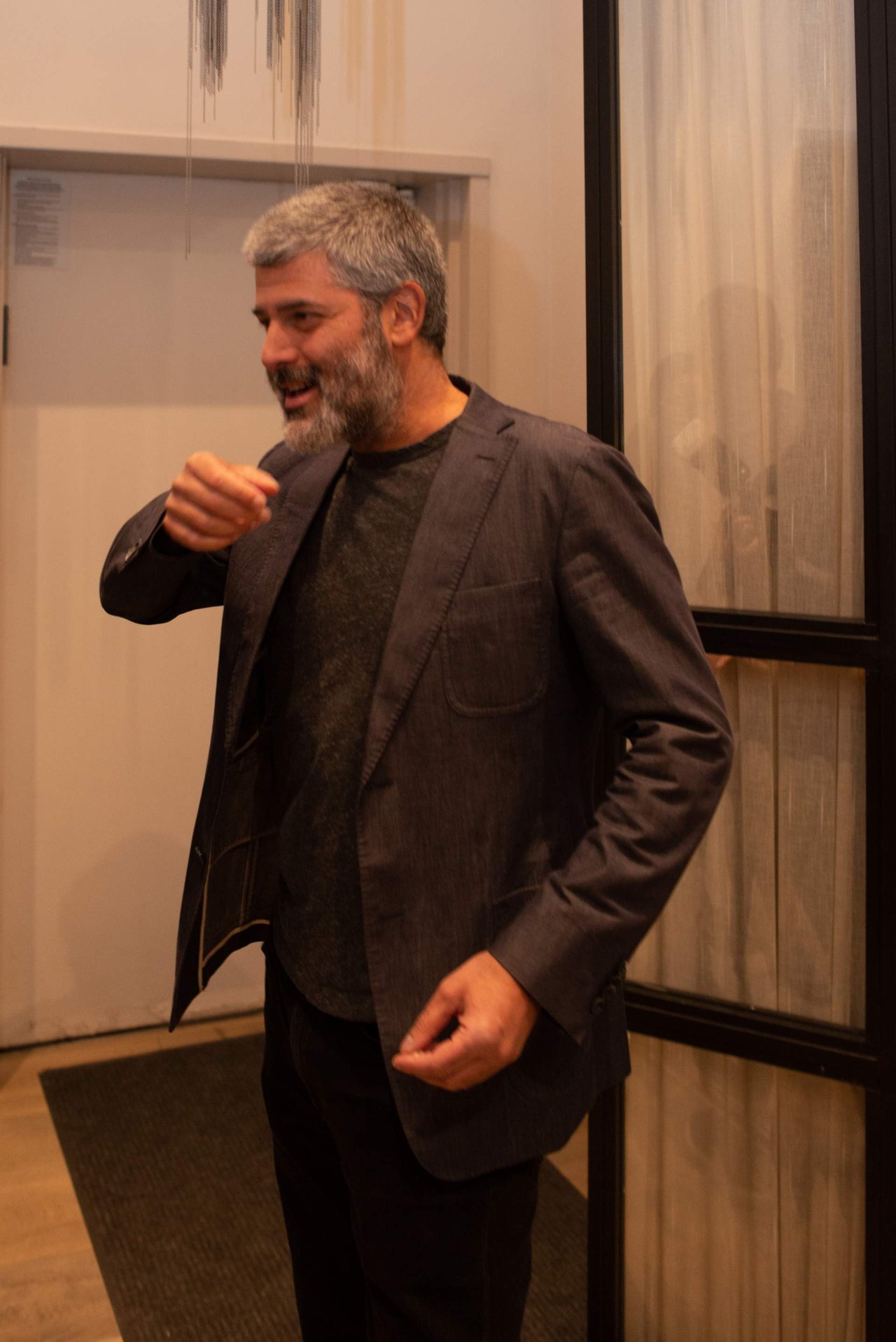 Gray-haired, bearded man in a dark blazer gesturing while speaking indoors.