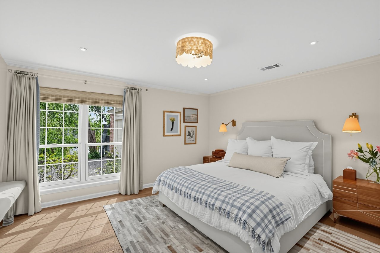 Bright bedroom with a white bed, gray plaid throw, and large window overlooking greenery.