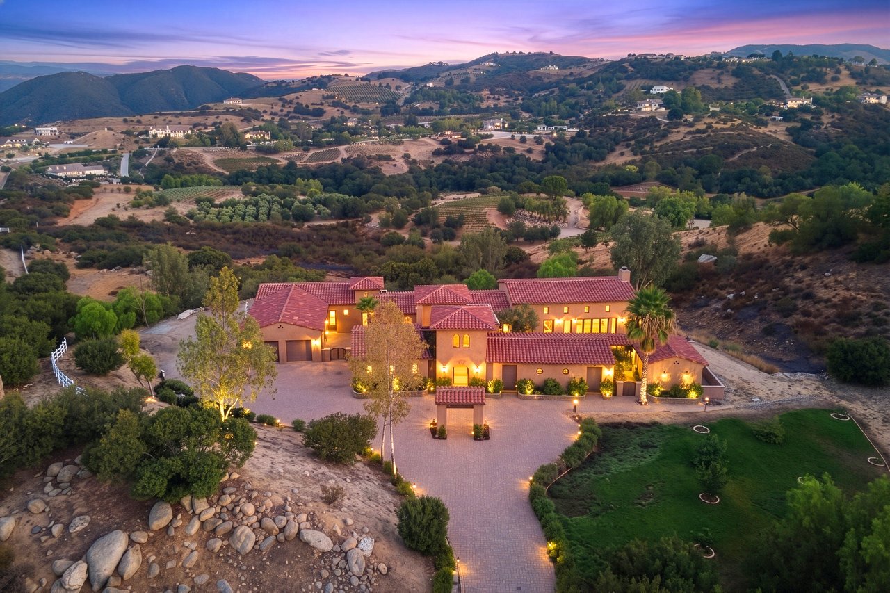 Aerial view of a red-tile-roof home and driveway beneath rolling hills at dusk.