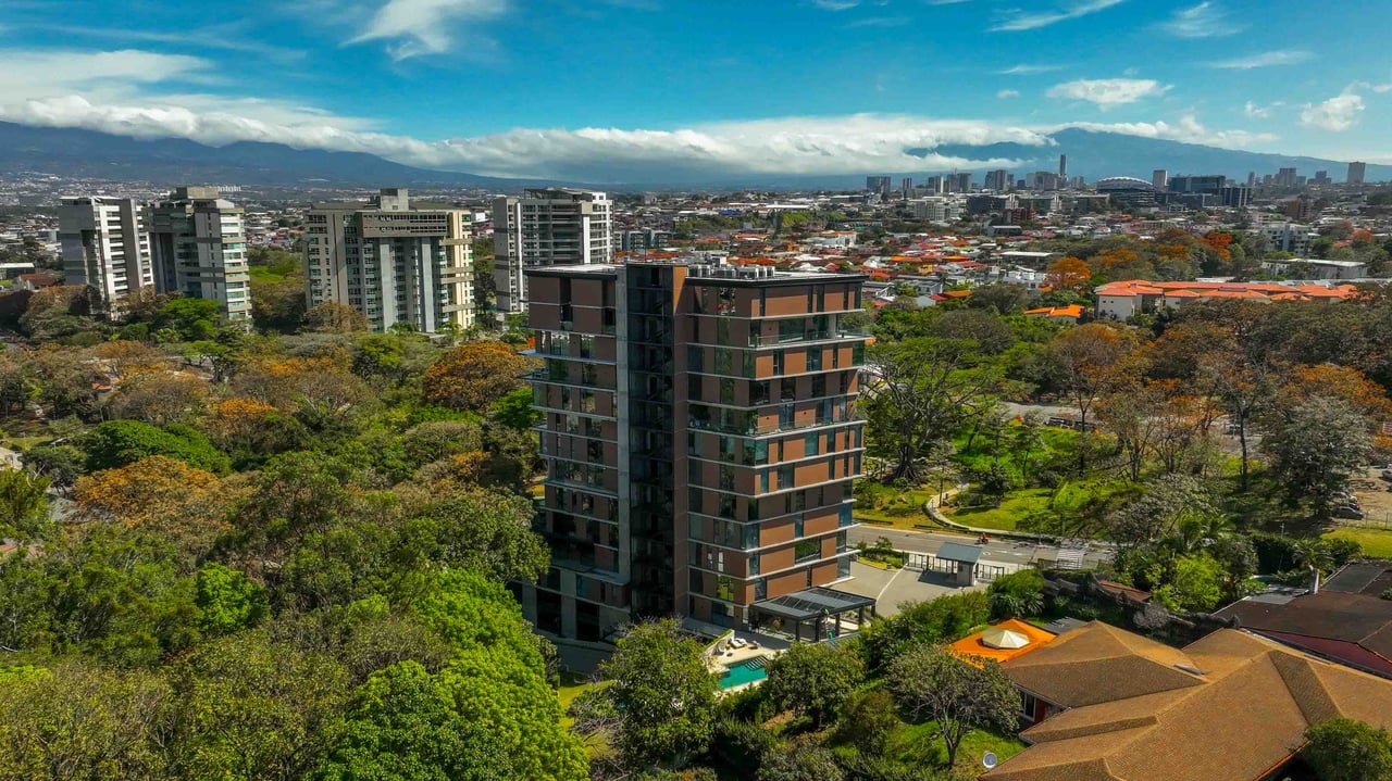 Modern apartment building surrounded by trees, with a city skyline and mountains in the distance.