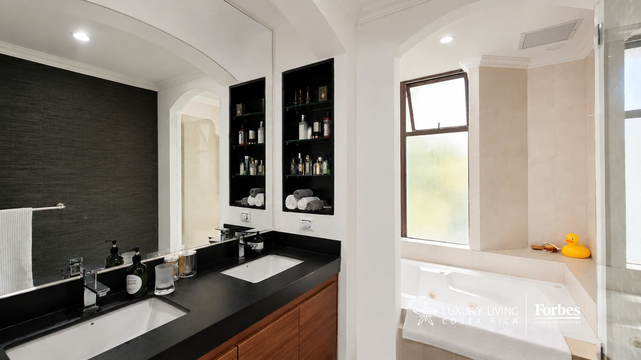 Condominium bathroom with a double vanity, black counters, wood cabinets, and a bathtub beneath a tall window.