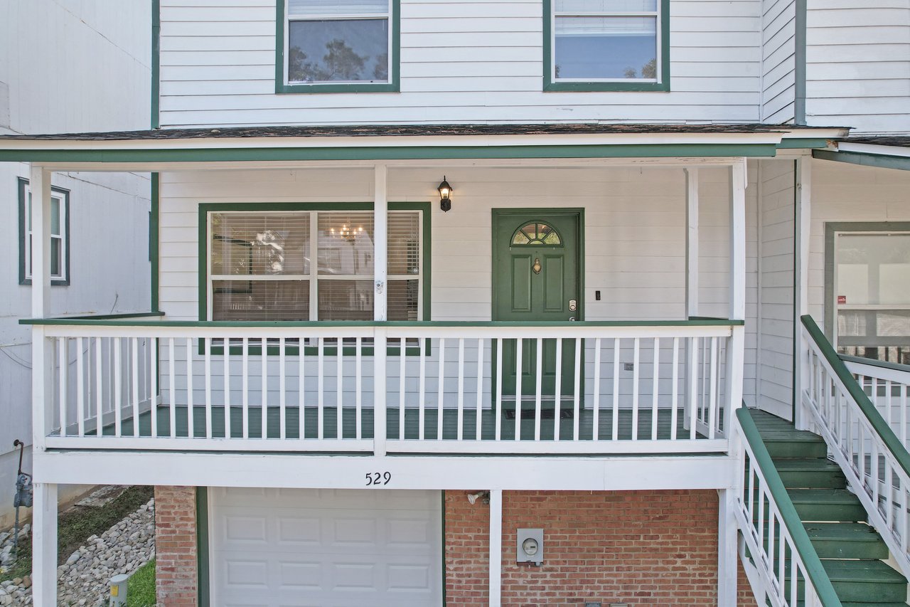 Front porch of the townhome at 529 Collinsford Road, Tallahassee, Florida, featuring a covered entry with white railings, green trim, and a matching green front door. A hanging porch light illuminates the entrance, and a large front window provides a view into the dining area. Brick accents frame the garage below.