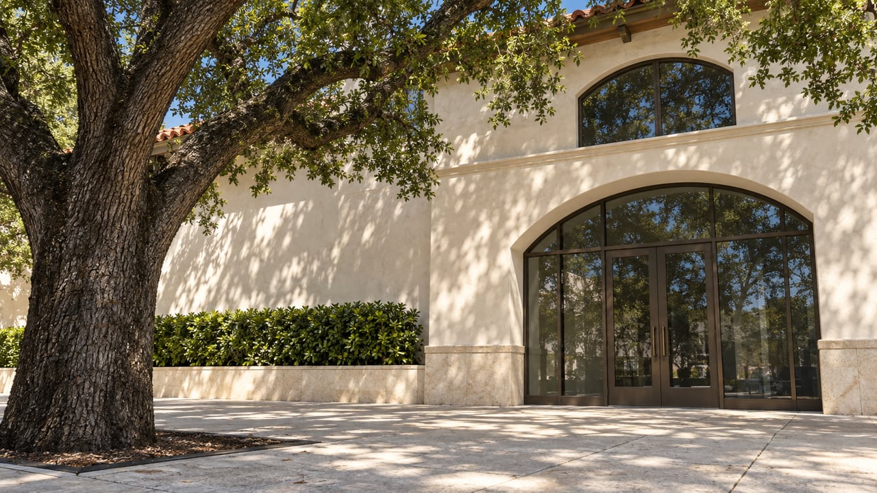 Live oak shading an ivory stucco storefront with an arched bronze-framed glass entrance.