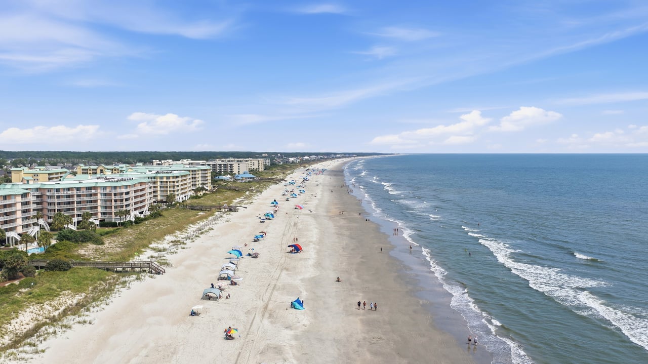 Aerial view of a sandy beach and coastal condominiums, with umbrellas along the shoreline.