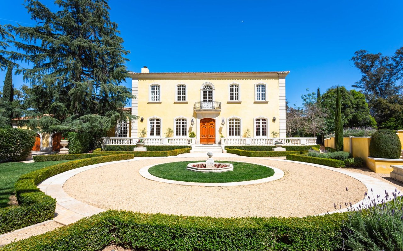 French-style estate with a circular gravel driveway, central fountain, clipped hedges, and orange front door.