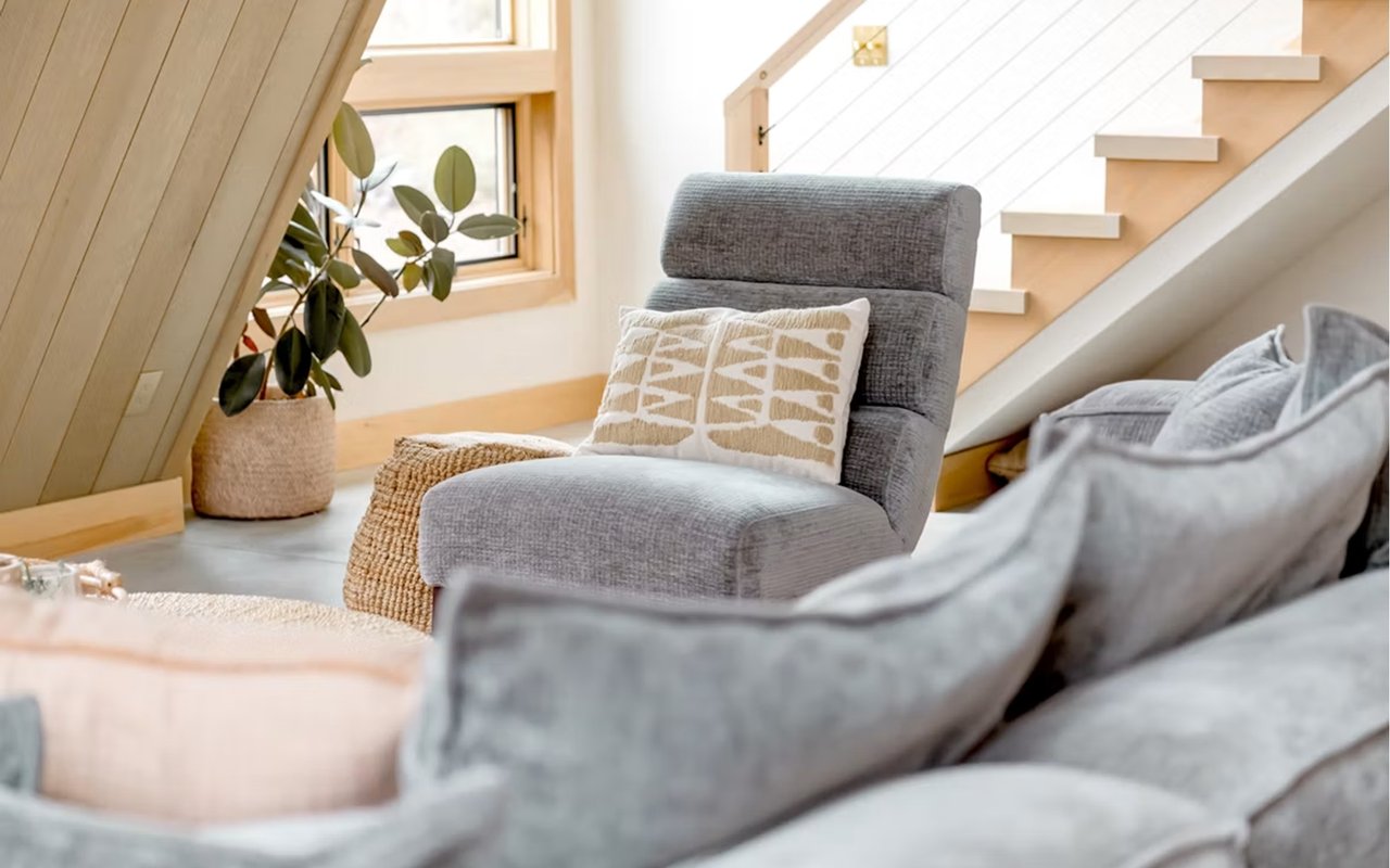 Staged living room with gray seating, a potted plant, and pale wood stairs.