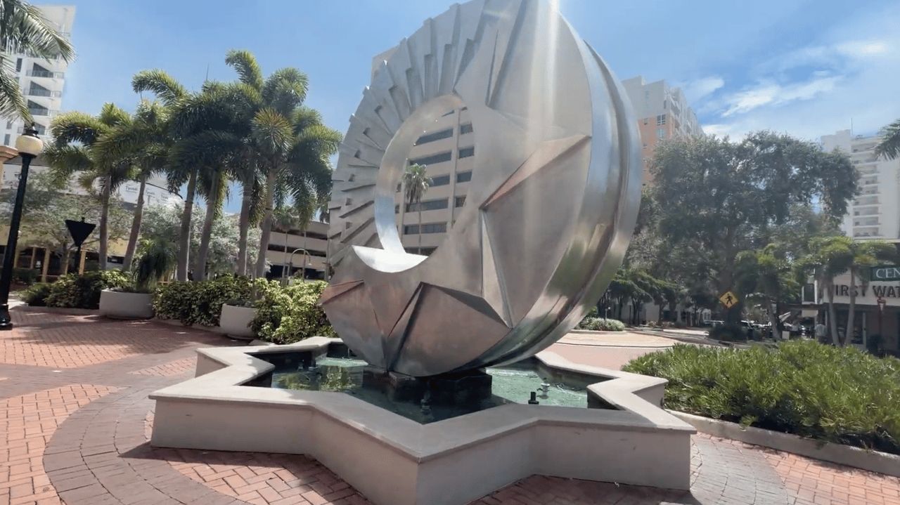 Downtown plaza with a large abstract metal sculpture fountain, brick paving, palm trees, and planters.
