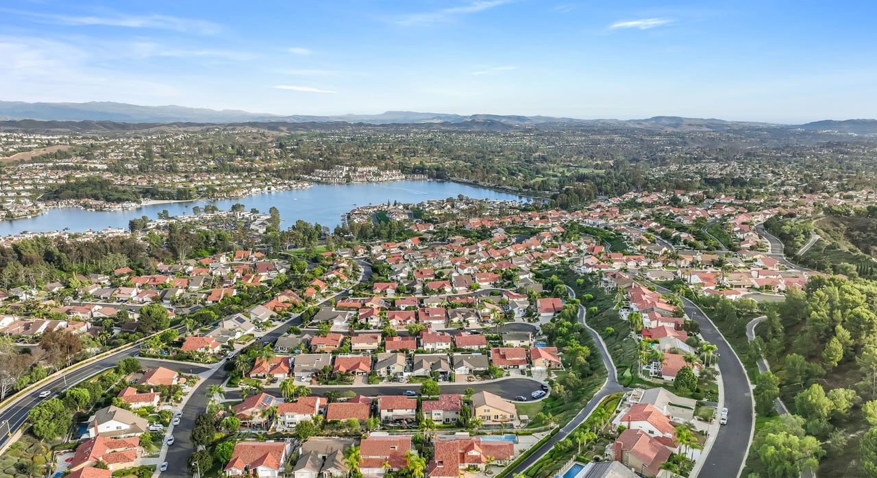 Aerial view of red-tile-roof homes and curving streets in Madrid del Lago, Mission Viejo, with Lake Mission Viejo and distant hills under a blue sky.