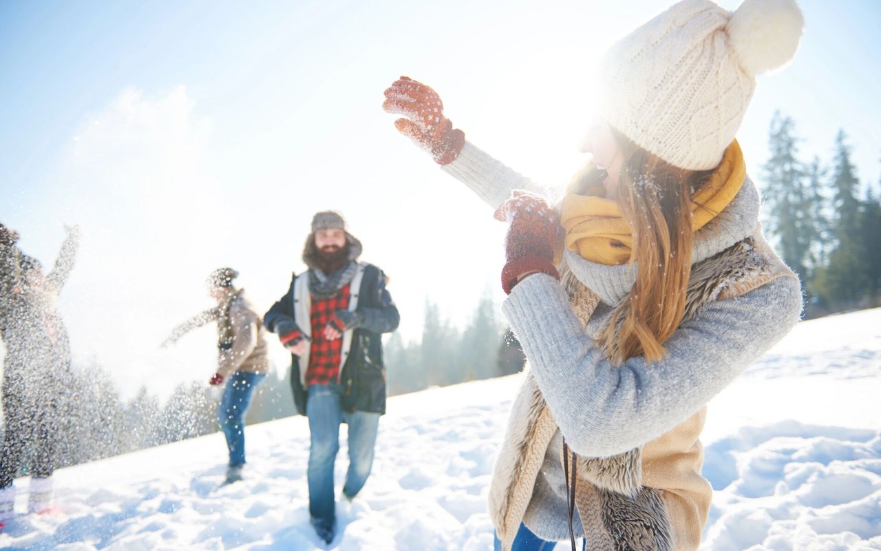People play in the snow; a woman in a white knit hat raises her arms in the foreground.