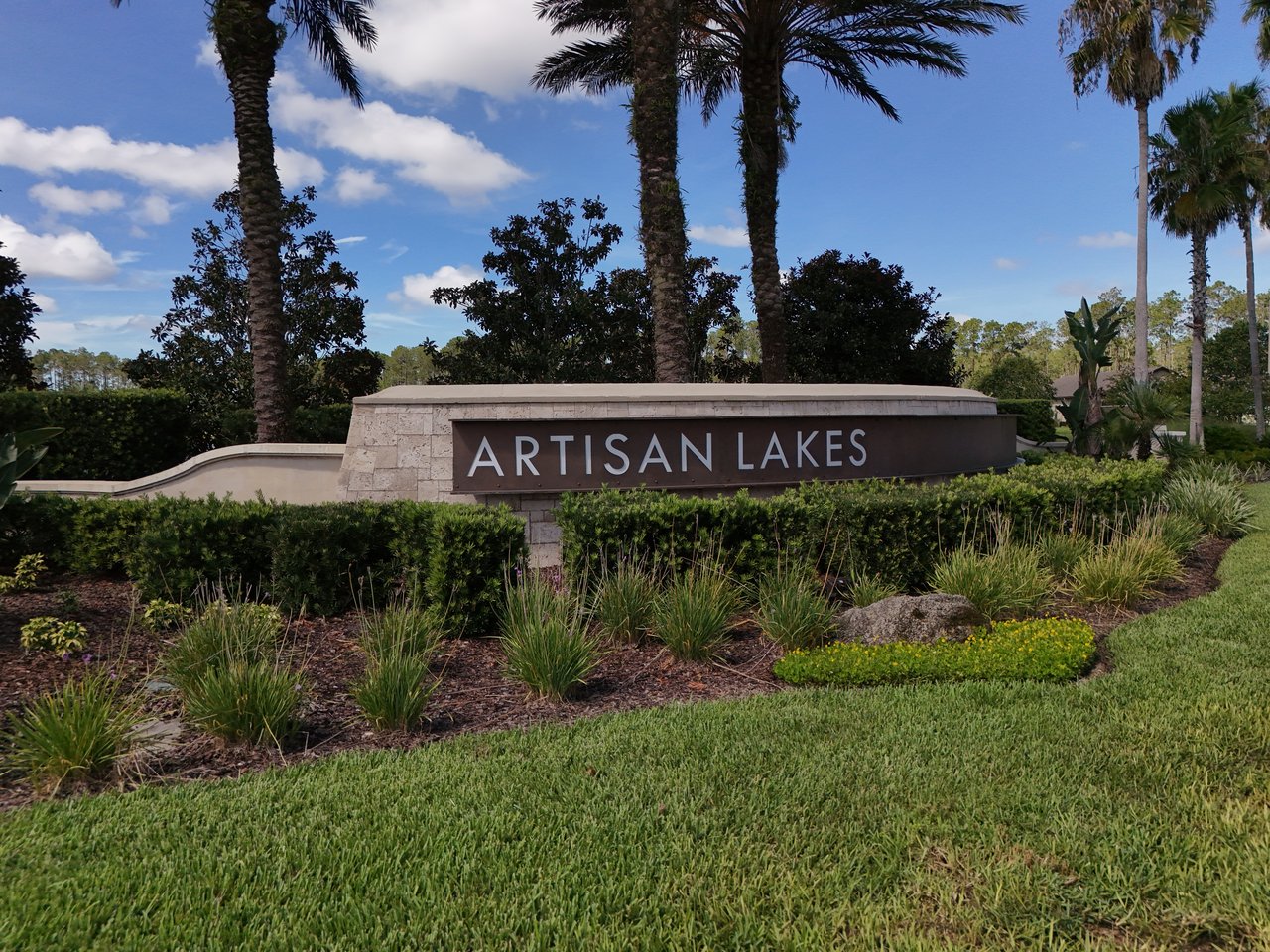 Artisan Lakes entrance sign among landscaped shrubs and palm trees.