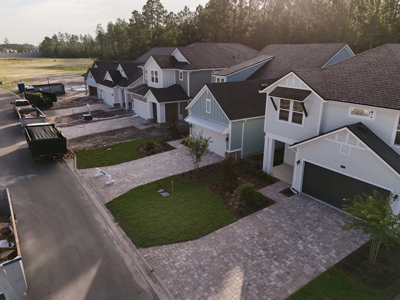 New homes with brick-paver driveways line a residential street beside a wooded tree line.