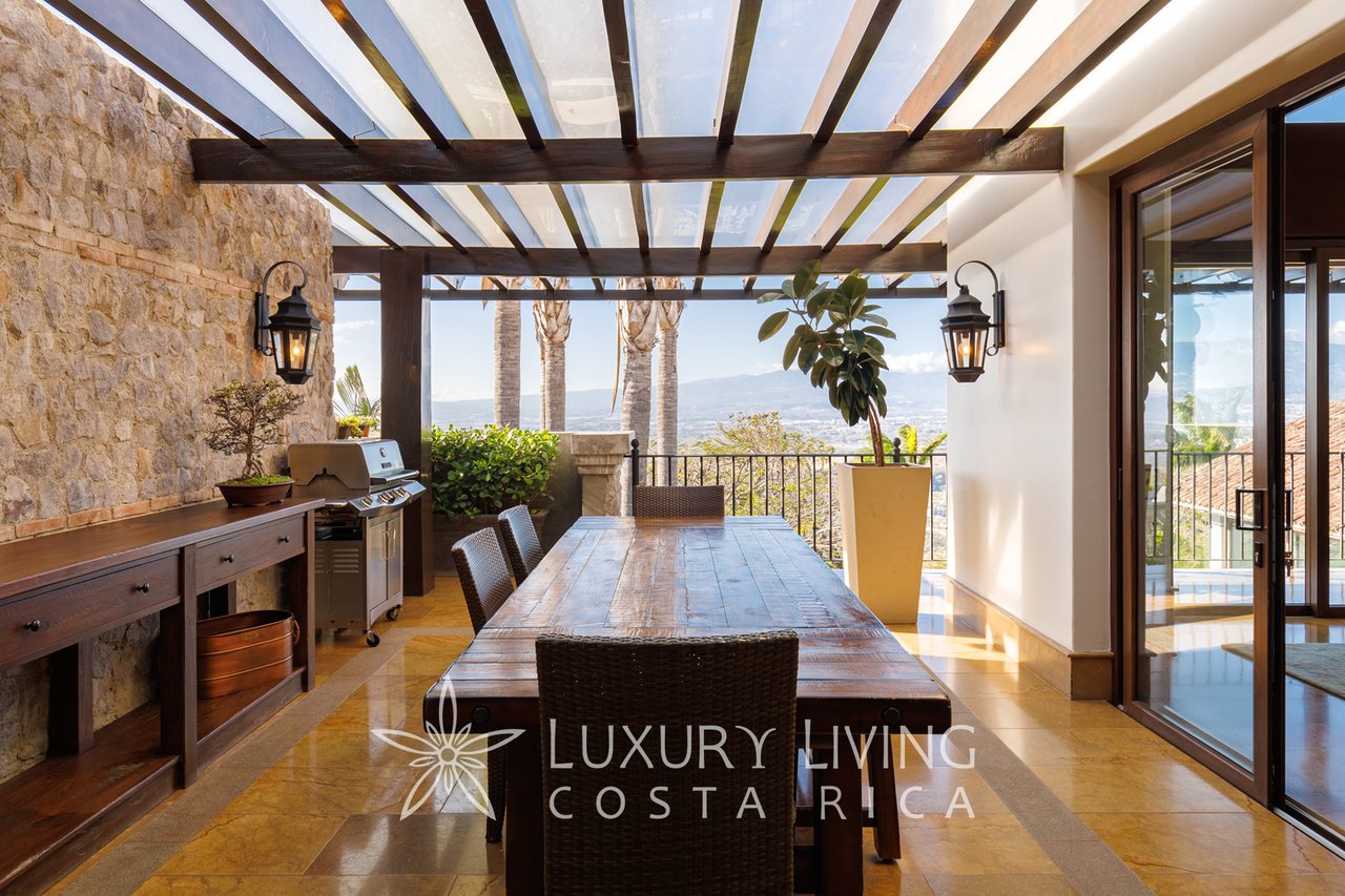 Covered terrace with a long wooden dining table, stone wall, barbecue, and distant valley view.