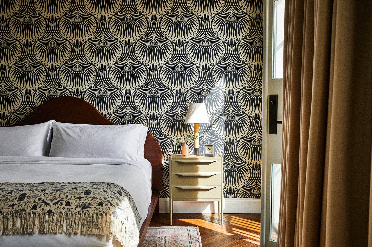 Bedroom with bold patterned wallpaper, a bedside chest, and sunlight across the hardwood floor.