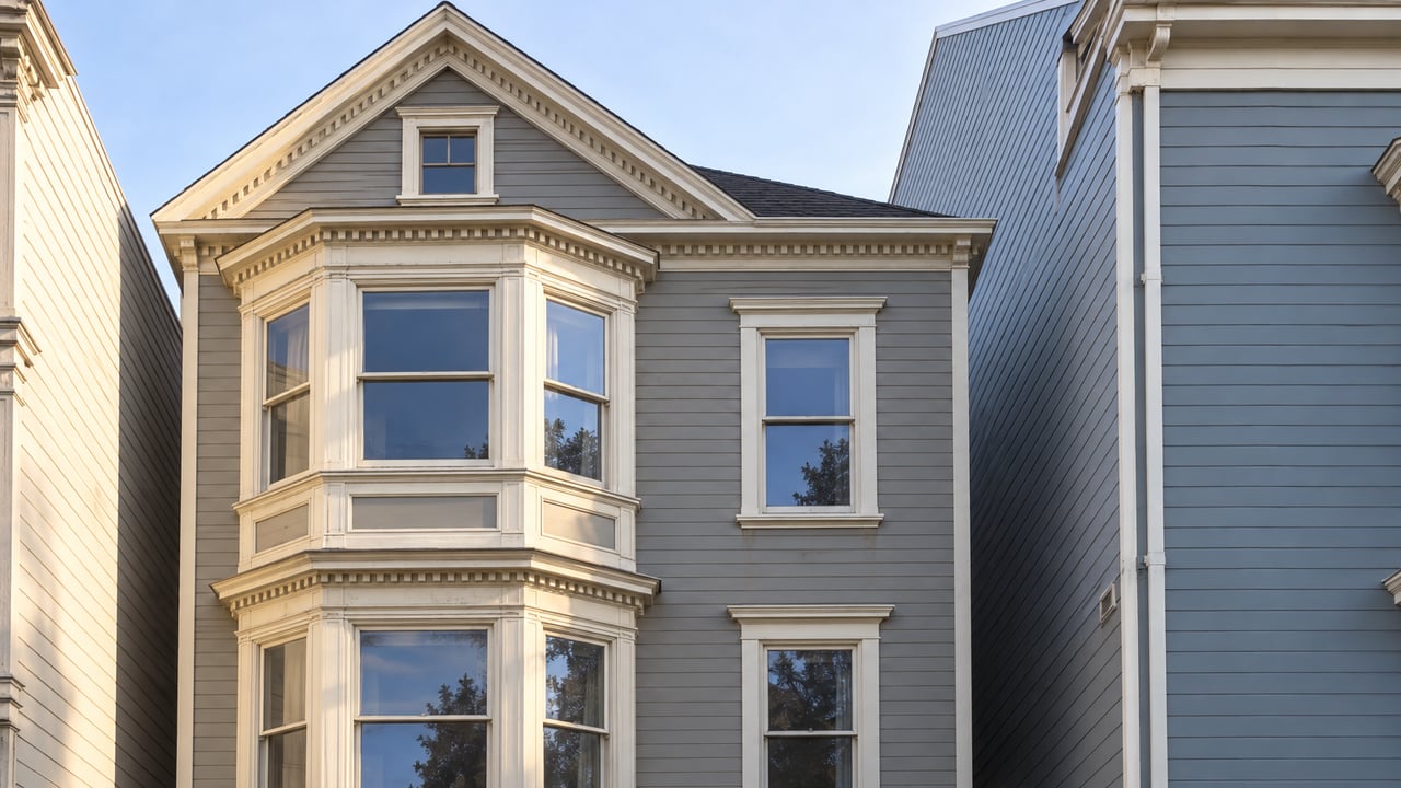 Three-story gray timber house with ivory bay-window trim, a raised oak entry and a camellia beside worn concrete steps.