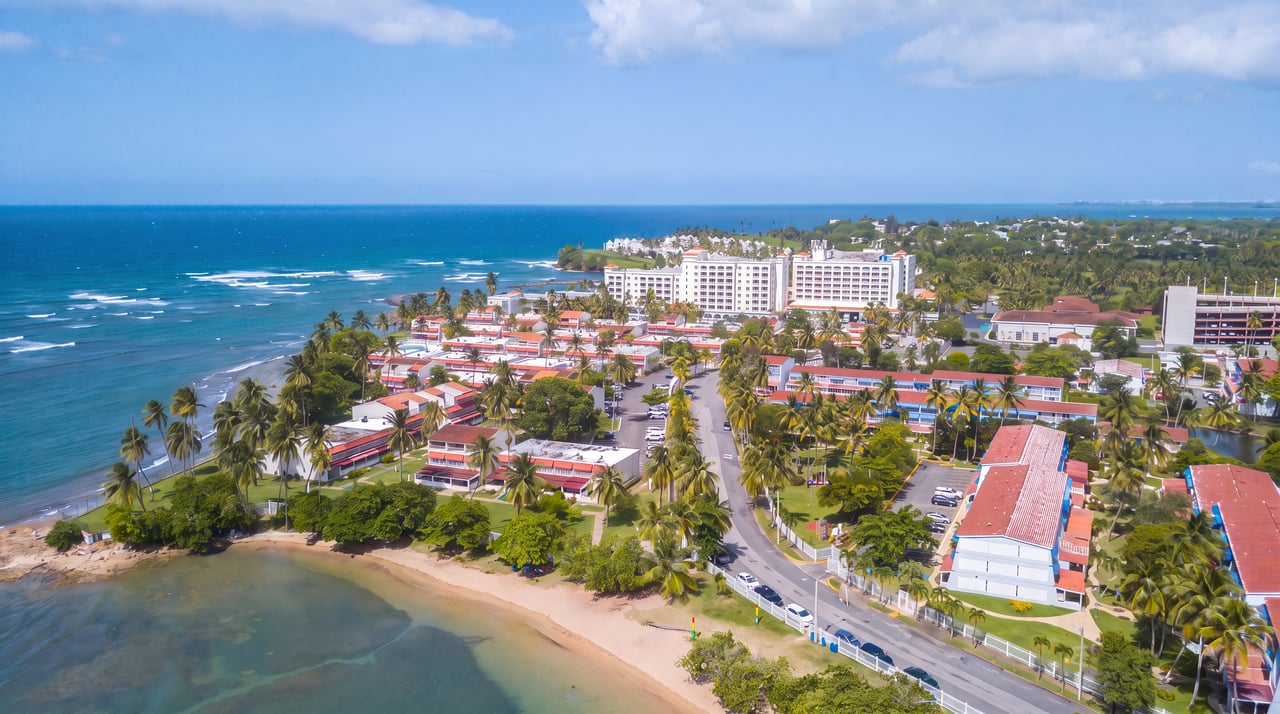 Aerial view of a palm-lined coastal development beside a sandy beach and turquoise ocean.