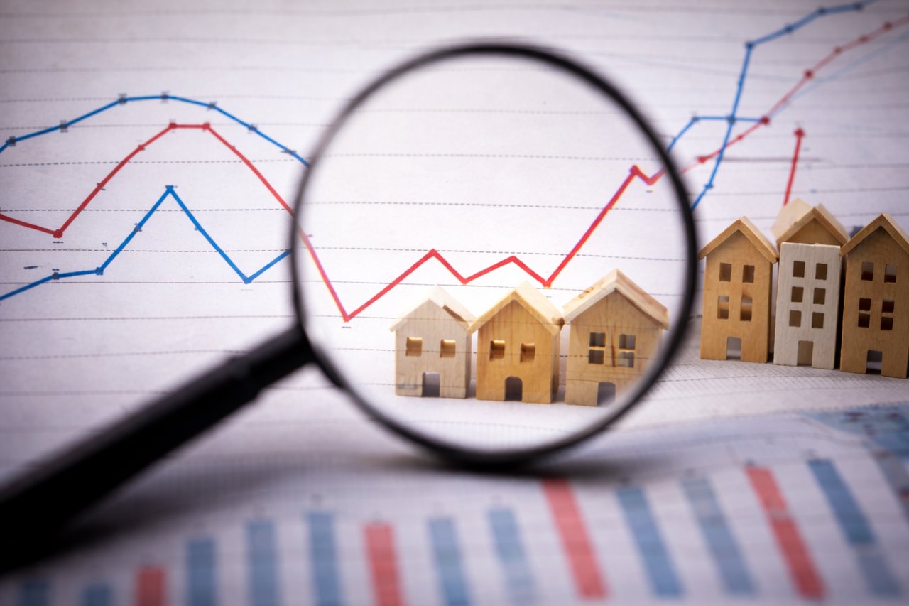 Wooden house models viewed through a magnifying glass with market charts showing changing home price trends