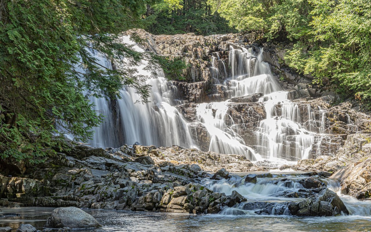 Multi-tiered waterfall flowing over rocky ledges into a forest stream.
