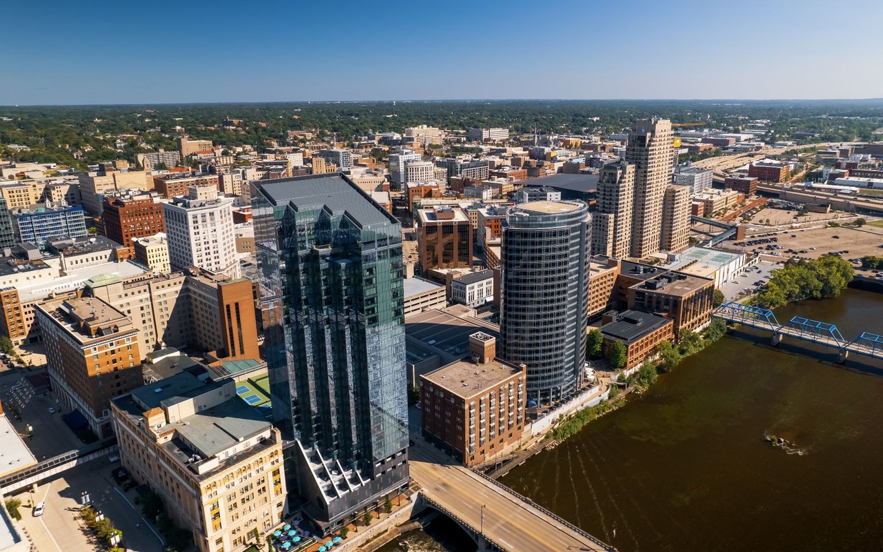 Aerial view of downtown buildings along a river, with a blue bridge and tree-lined horizon.
