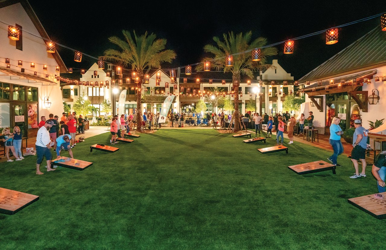 People play cornhole in a palm-lined courtyard with restaurants and string lights at night.