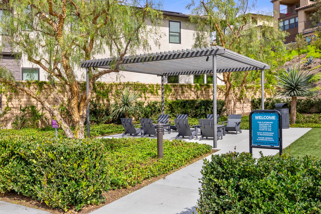 Outdoor community seating area beneath a pergola, with landscaped paths and a playground rules sign.