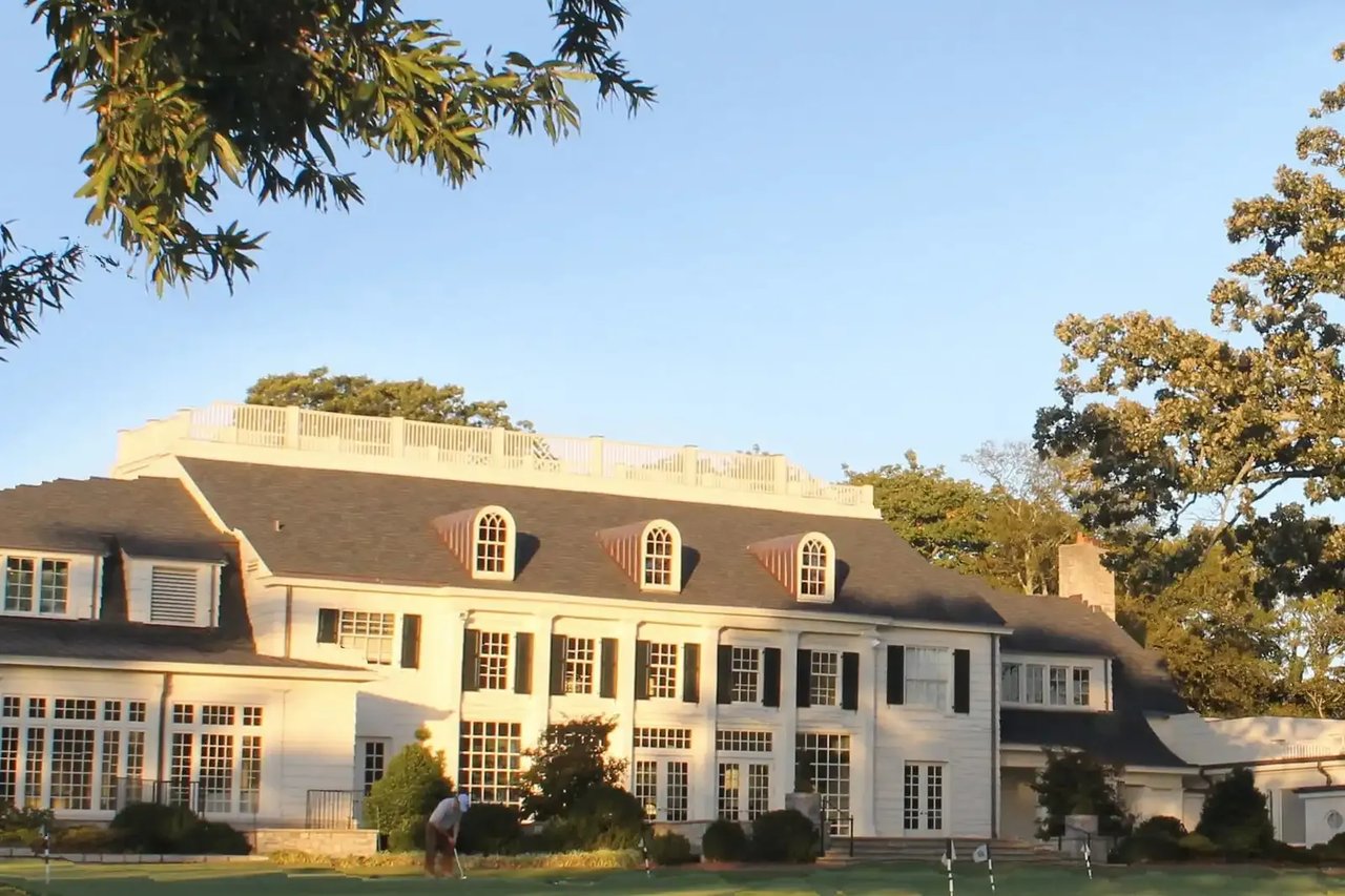Large white house with a rooftop terrace overlooking a lawn where a golfer putts.