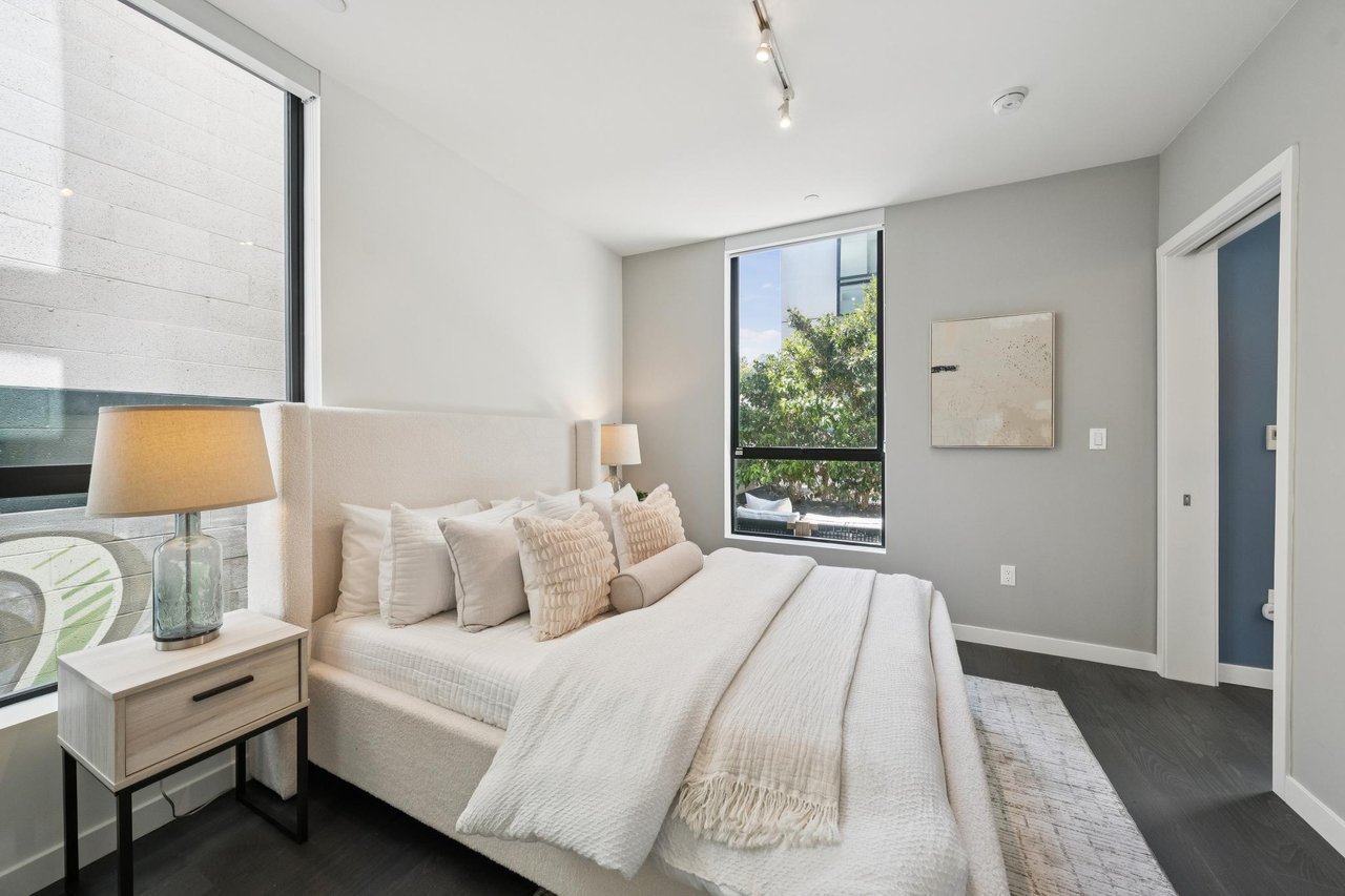 Staged bedroom with a white upholstered bed and tall windows overlooking greenery.