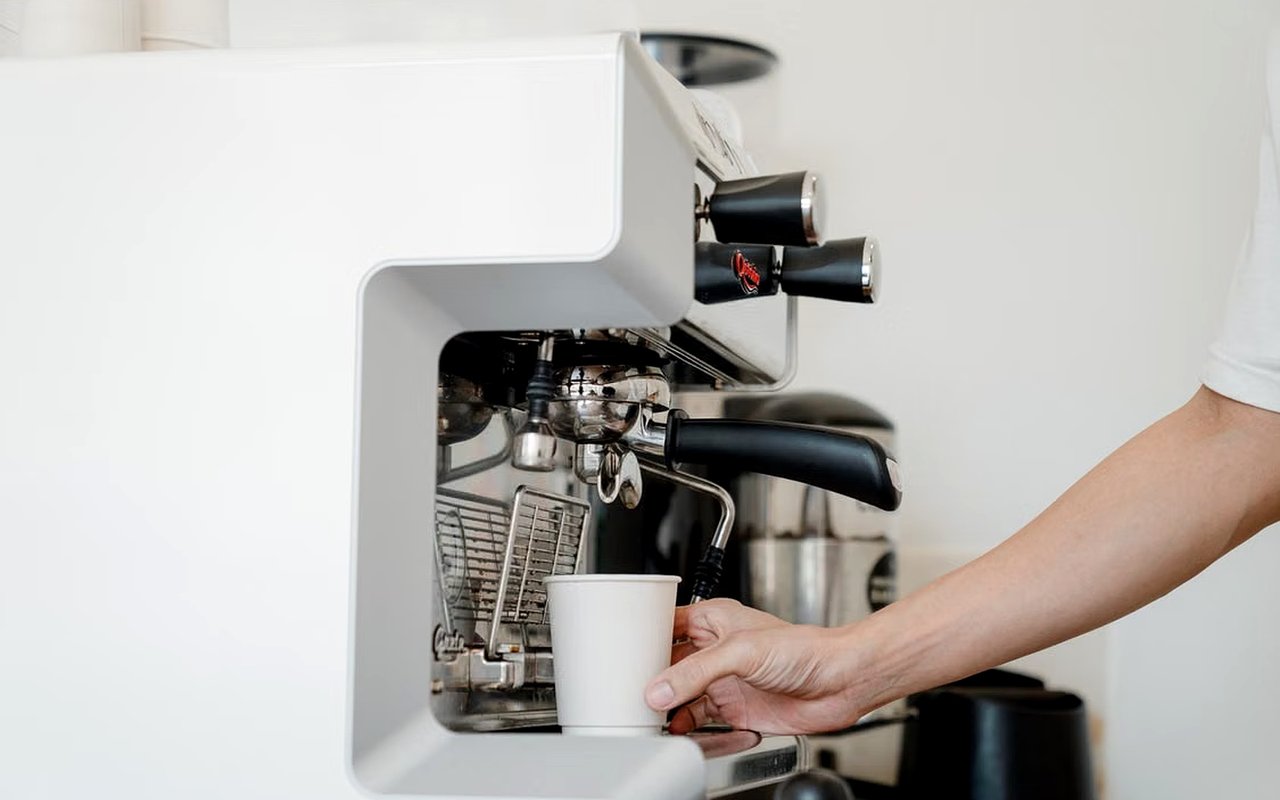 A hand places a paper cup beneath a large espresso machine.