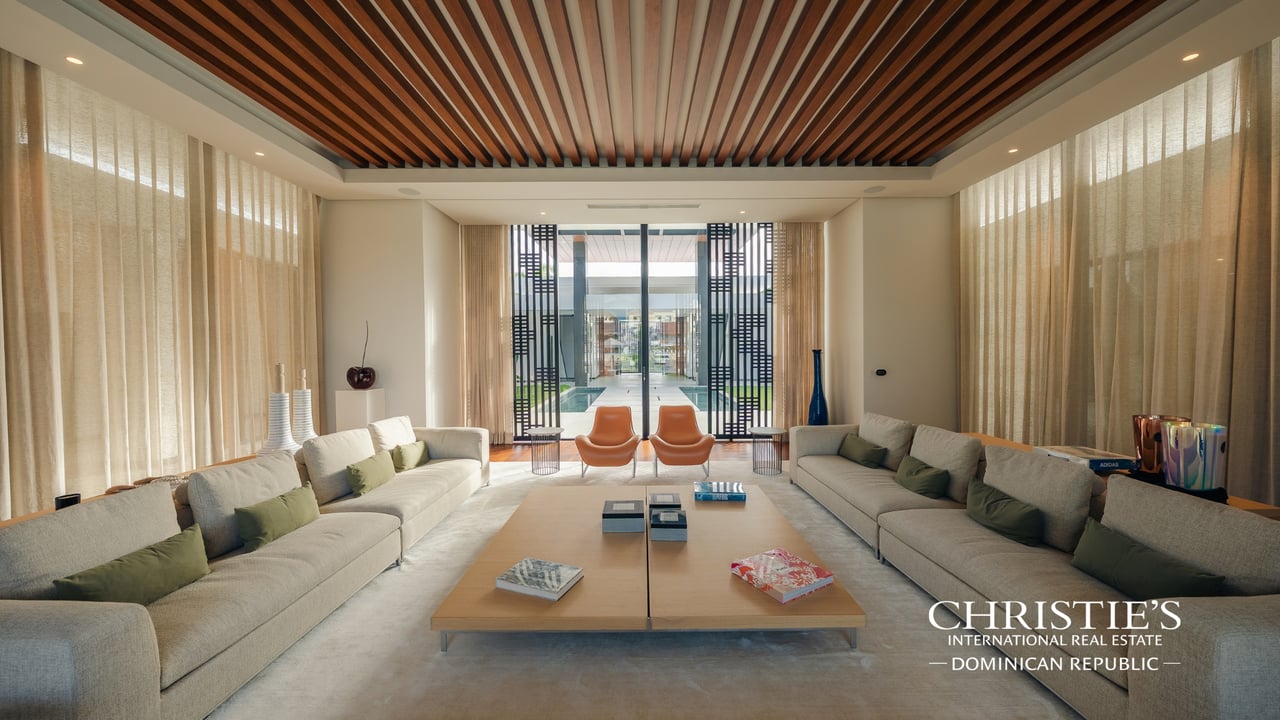 Villa living room with twin sofas, orange chairs, and a wood-slat ceiling facing a glass-walled courtyard.