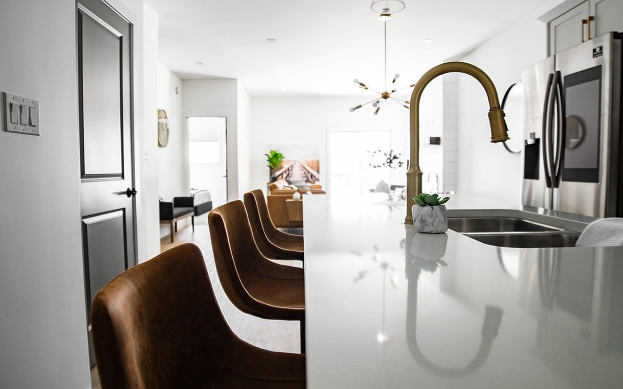 Modern white kitchen with a long island, brass faucet, stainless steel refrigerator, and brown counter stools.