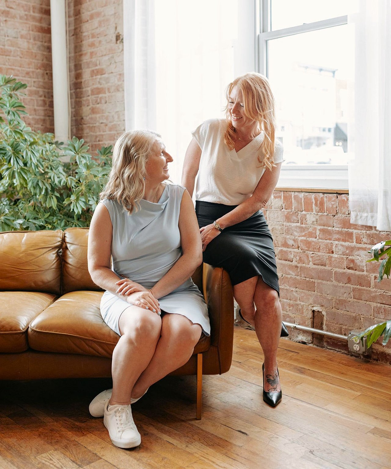Two women sit together in a brick-walled office, smiling at each other.