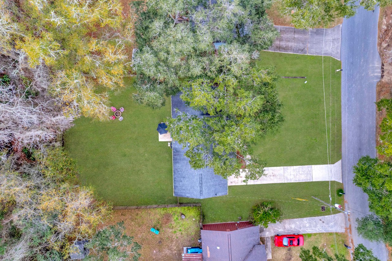 Aerial view of a suburban house with a gray roof, surrounded by lush trees and a large grassy yard. A red car is parked in the driveway, and a circular seating area with chairs is visible in the backyard.