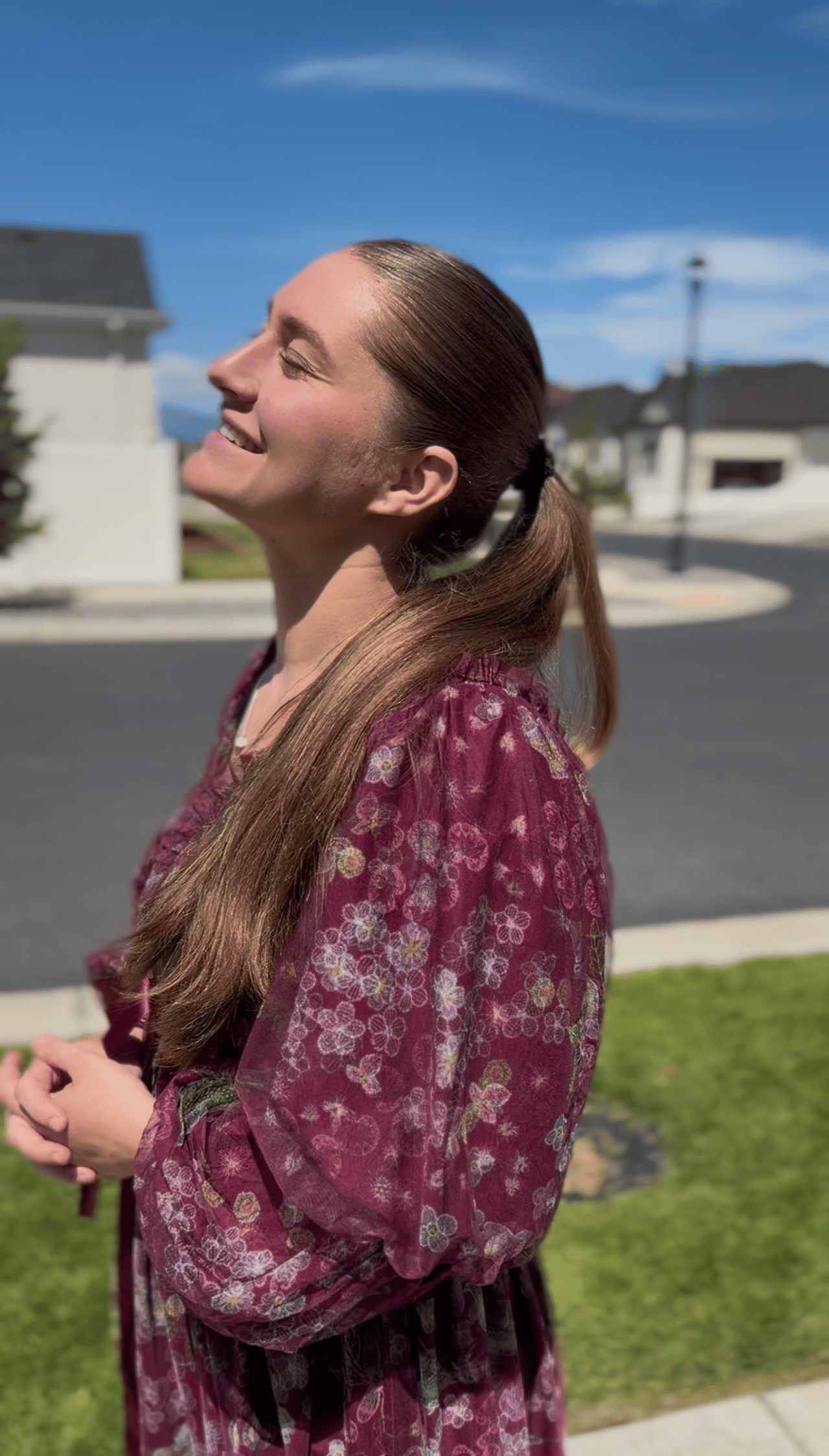 A woman smiling with eyes closed in natural sunlight outside a Utah neighborhood on a clear day