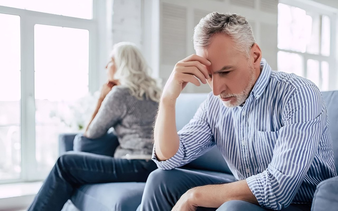 Distressed couple sitting apart on a sofa, man leaning forward with his hand on his forehead.