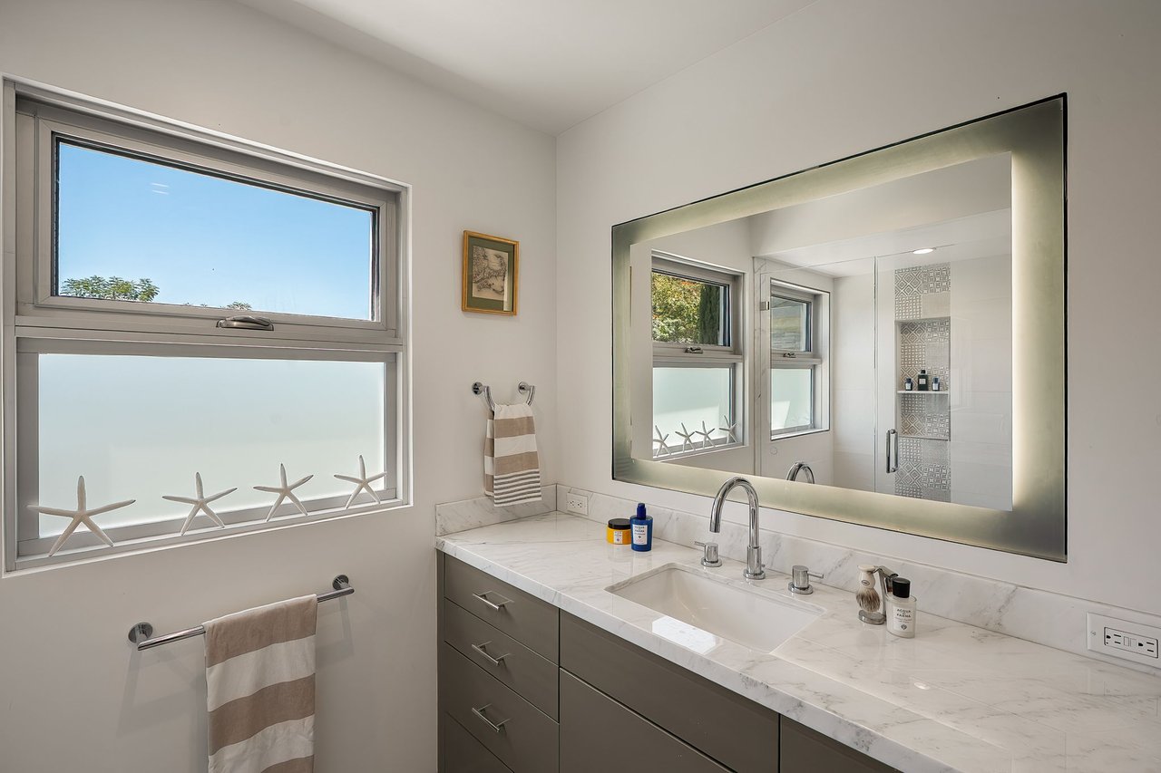 Bathroom vanity with marble countertop, gray cabinets, illuminated mirror, and starfish decorations on the windowsill.