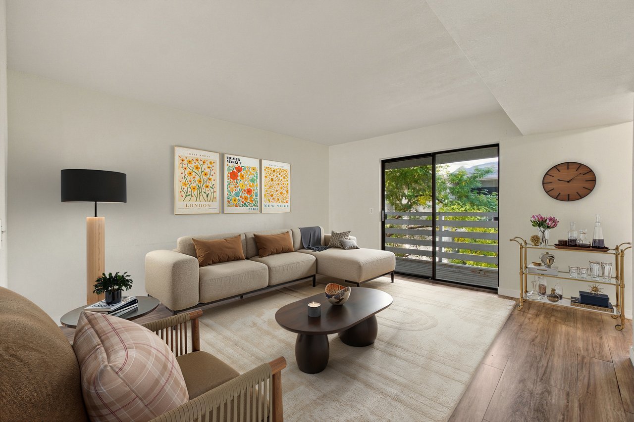 Living room with a beige sectional, floral city prints, and a sliding glass door to a leafy balcony.
