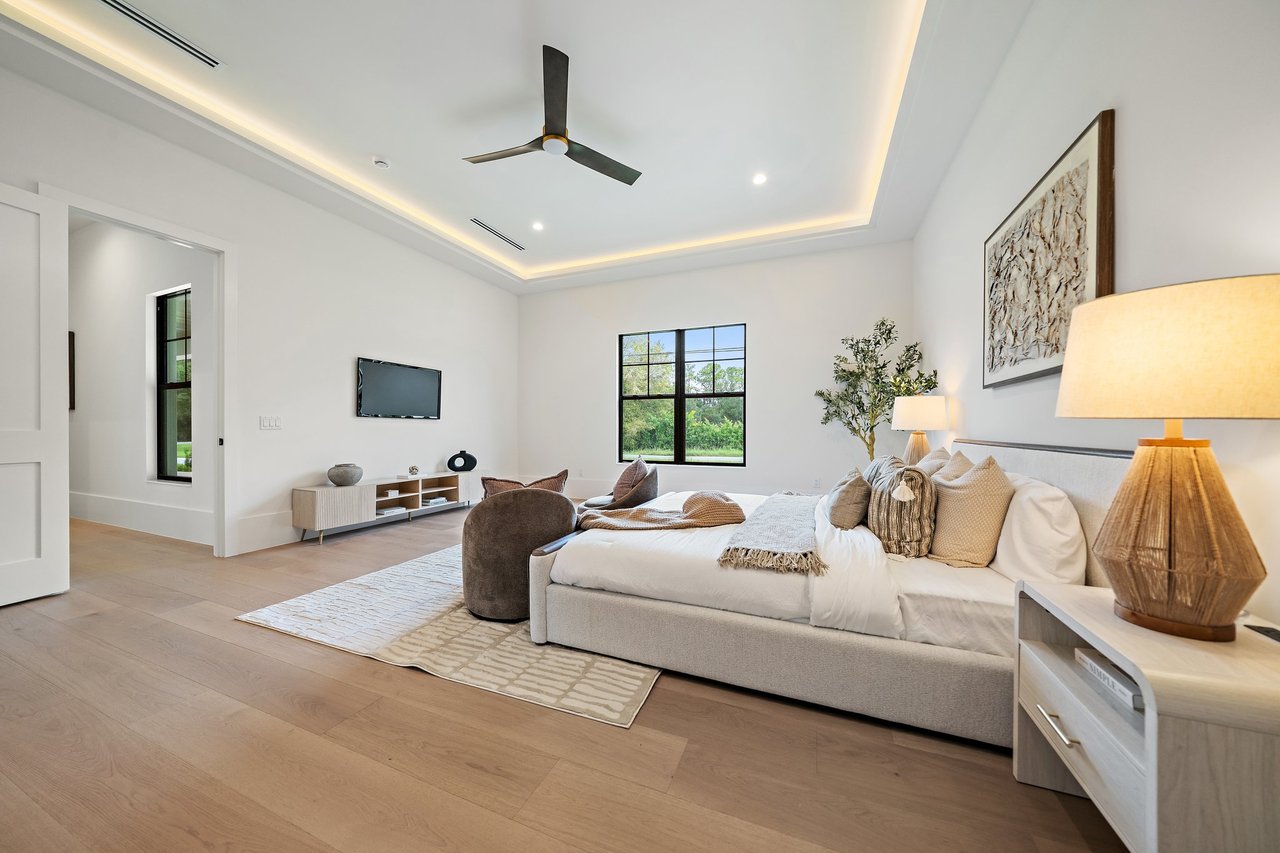 Staged bedroom with a large bed, tray ceiling, dark ceiling fan, and black-framed window.