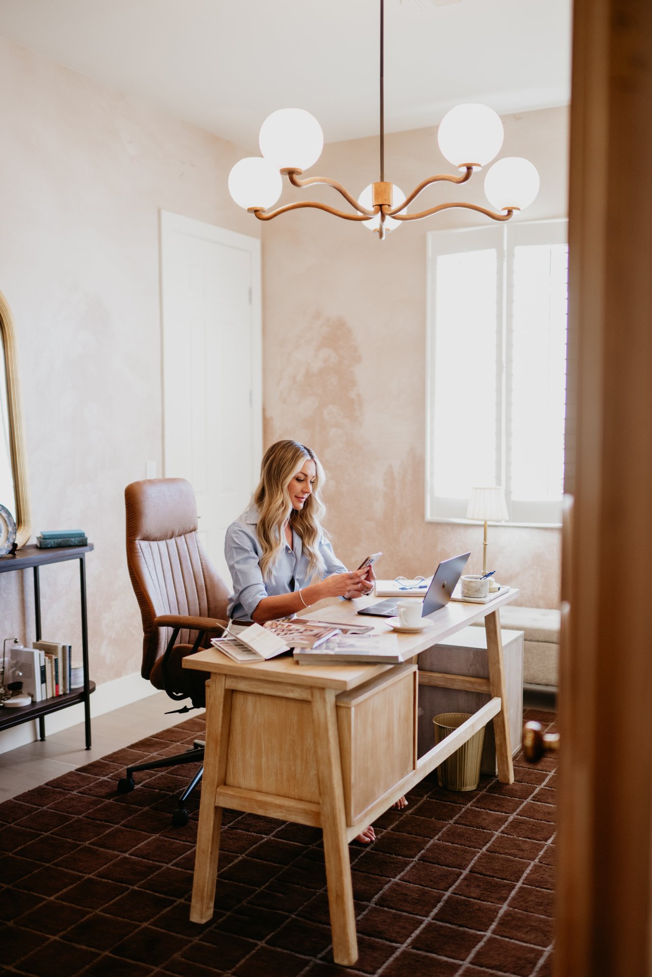 Kaylee Gallagher seated at a home-office desk, checking her phone beside an open laptop.